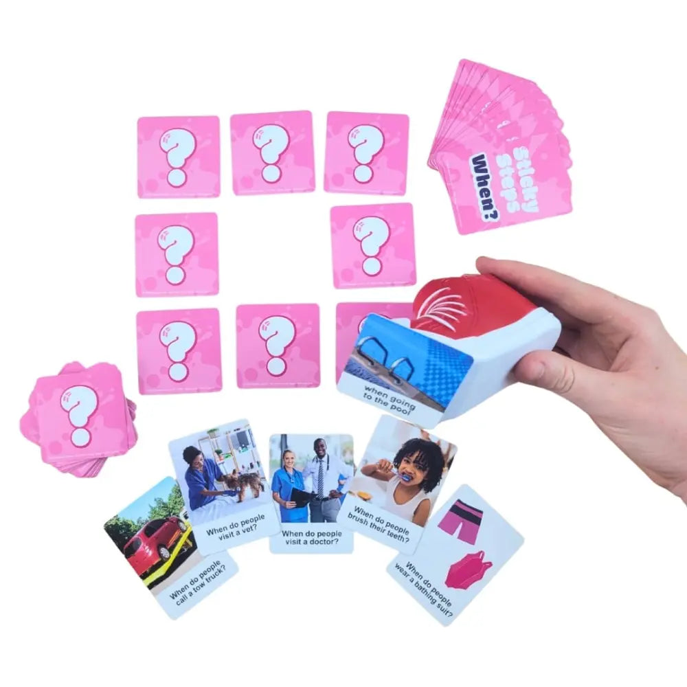 A hand holds a red and white card-picking device over a spread of pink question cards and illustrated cards on a white background.