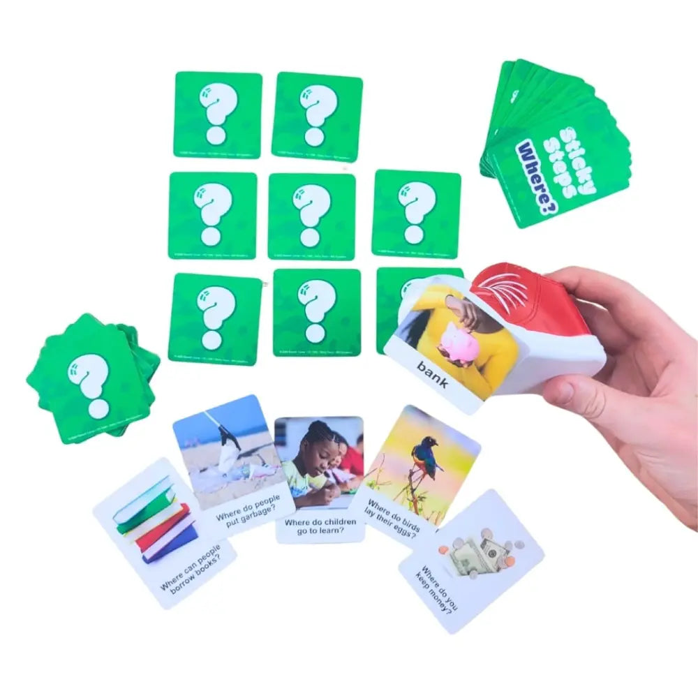 A children's language-learning card game set on a white background, featuring green question-mark cards, green 'Where is' cards, and white image-based cards being held by a hand.