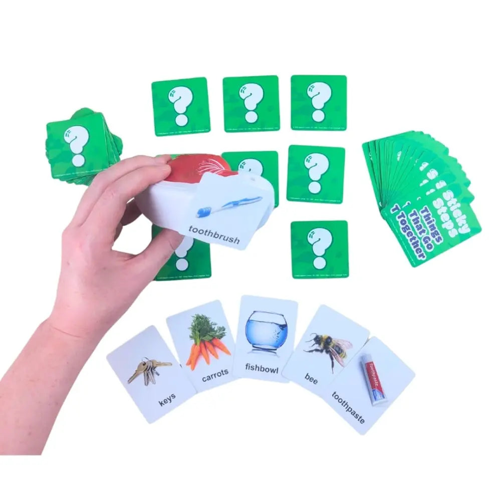 A children's educational card game set featuring green question cards and white illustrated cards, with a hand holding a toothbrush card. The set includes cards for keys, carrots, fishbowl, bee, and toothpaste, along with a deck of green cards and a stack of question cards on a white background.